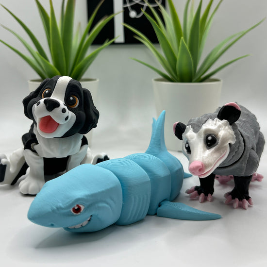 Three toy figures, one resembling a shark, one a opossum, and the other a dog, on a white surface with plants.