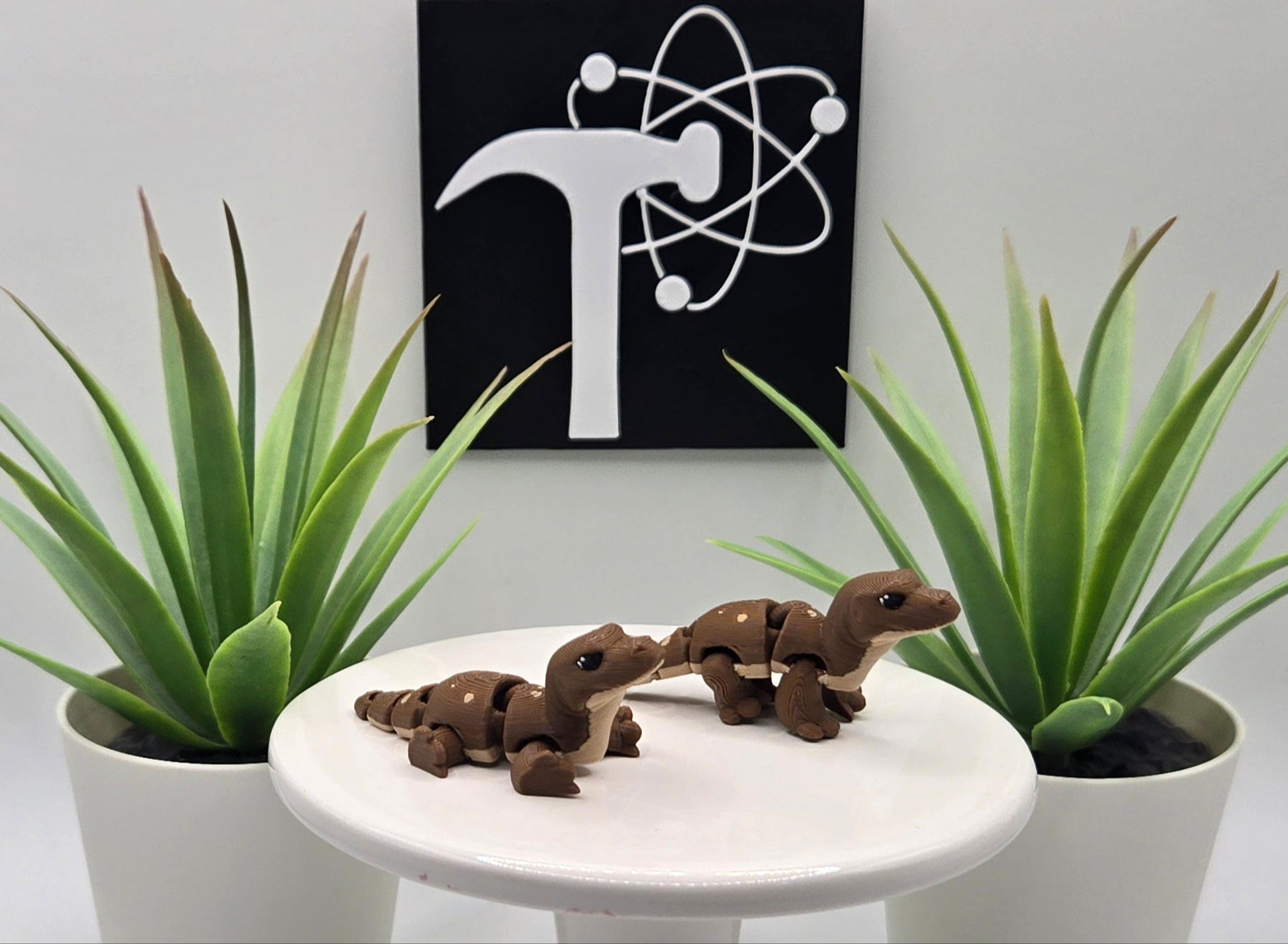 Two mini komodo dragon flexi animals on a white pedestal with plants and the Crafticist logo in the background.