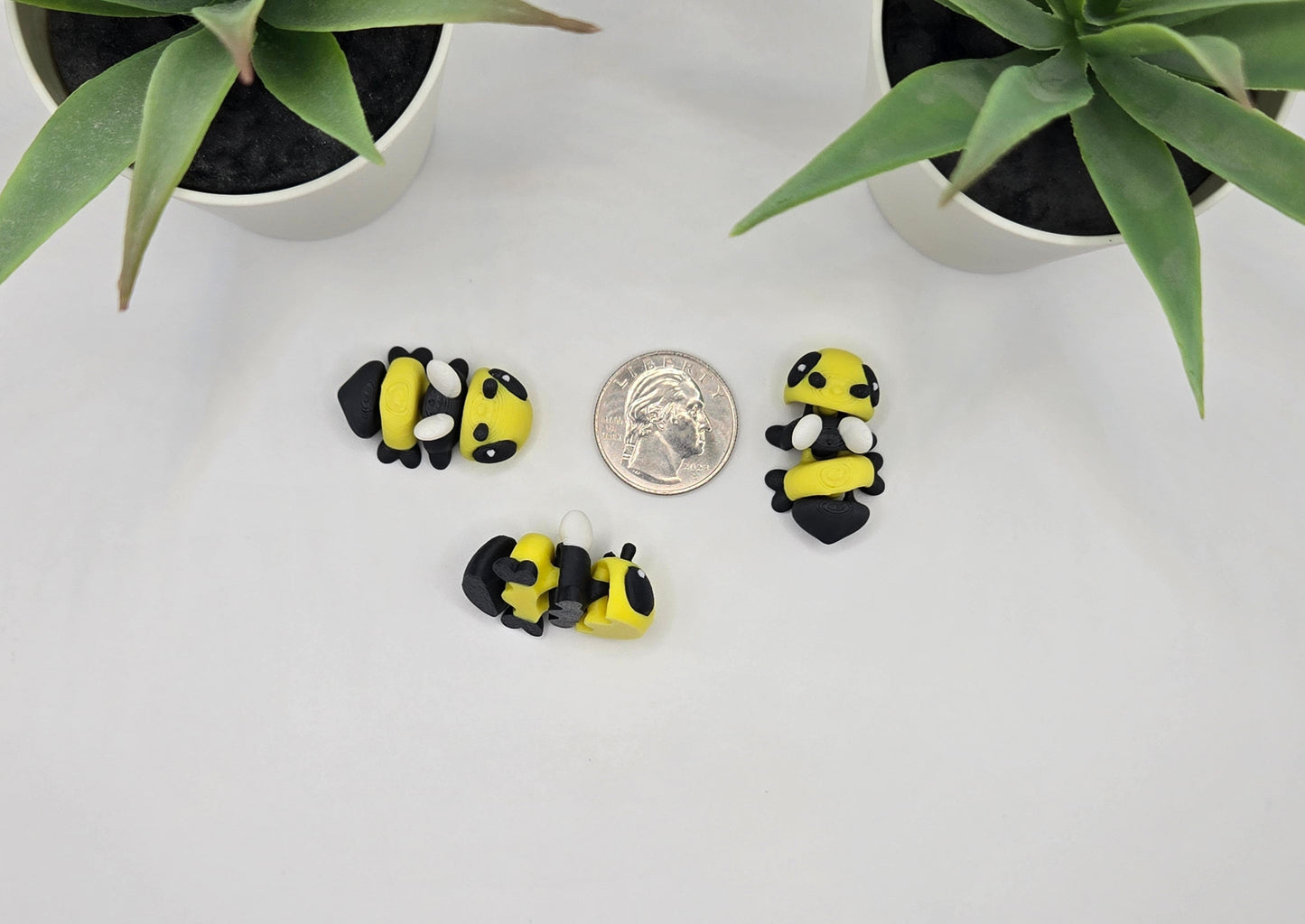 Three small bee toy figurines on a white surface with two potted plants in the background.