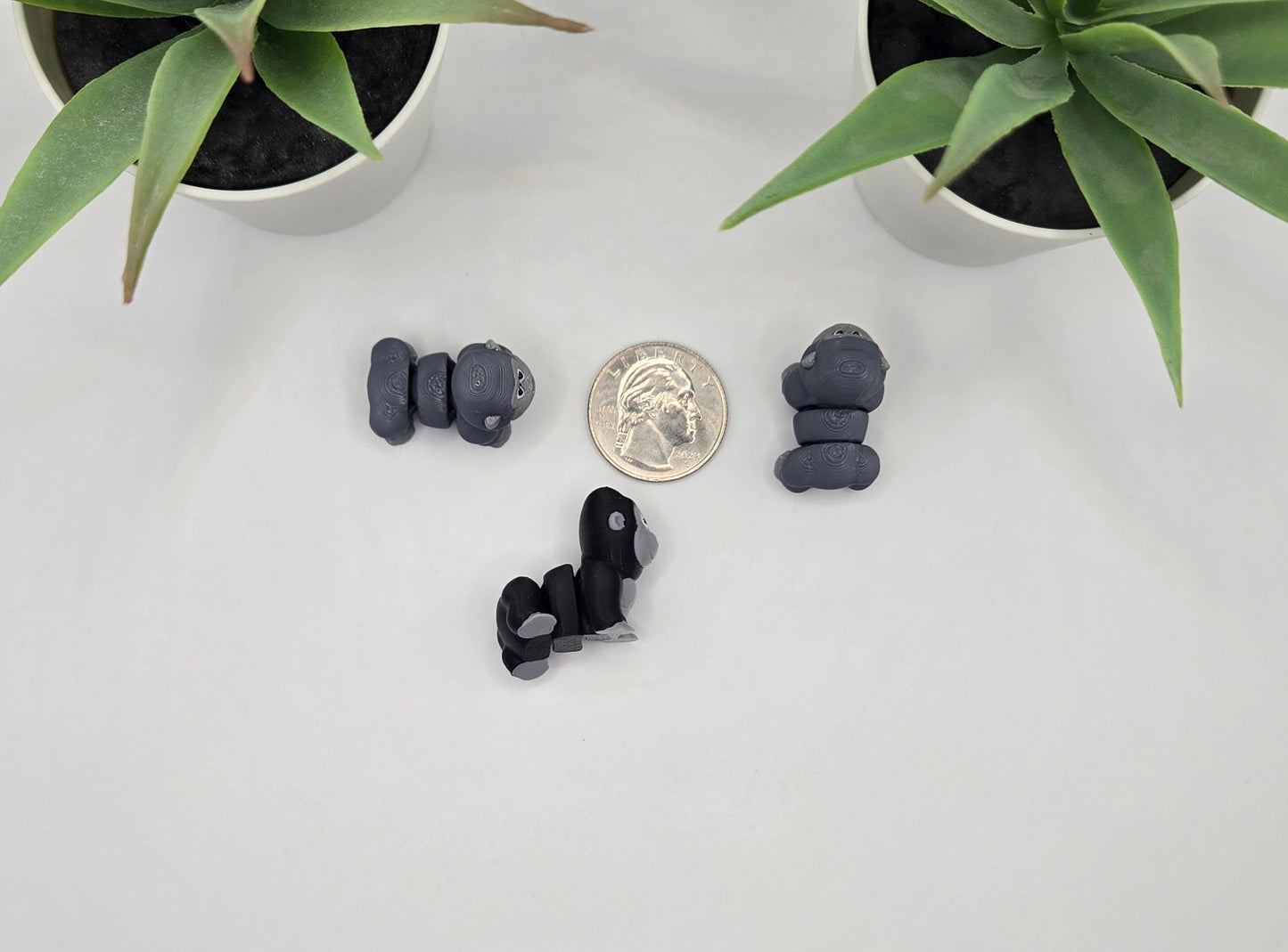 three small articulated gorilla toys with a quarter on a white surface with plants in the background