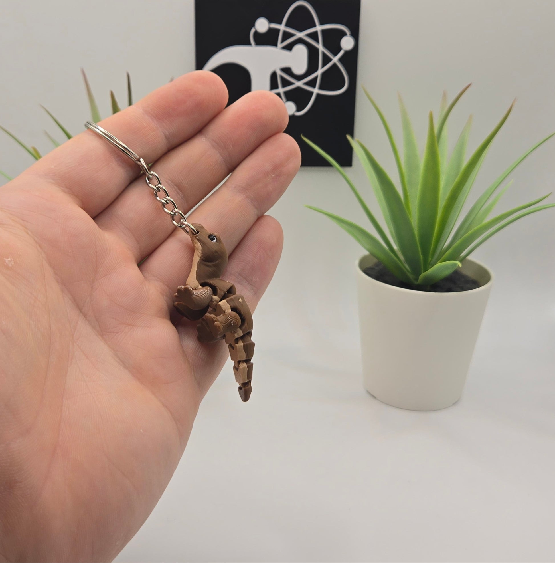 hand holding a small komodo dragon on a keychain with plants and the crafticist logo in the background