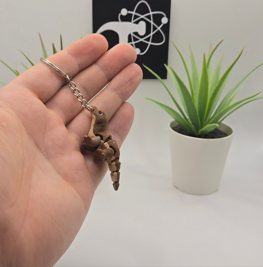 hand holding a small komodo dragon on a keychain with plants and the crafticist logo in the background