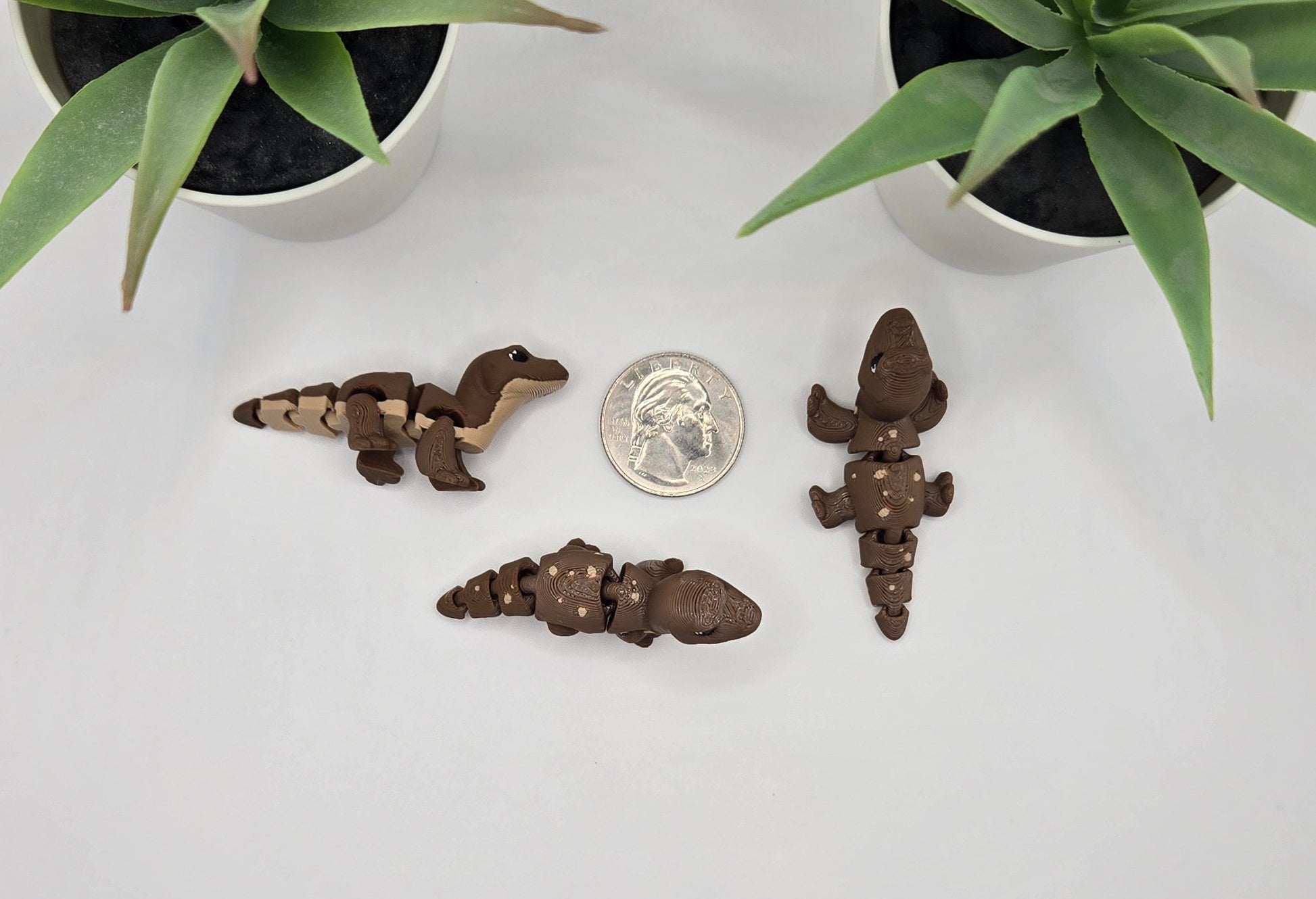 Three small komodo dragon figurines and a quarter on a white surface with plants in the background