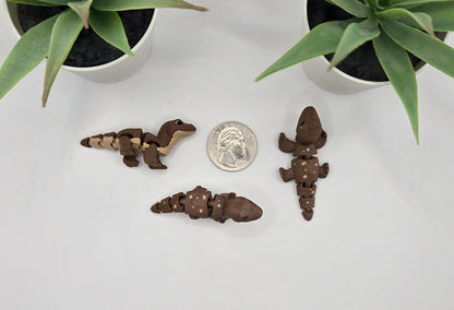 Three small komodo dragon figurines and a quarter on a white surface with plants in the background
